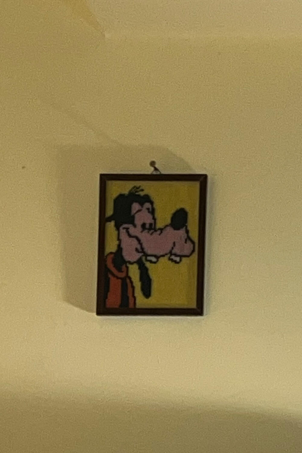 An einer beigen Wand hängt ein gerahmtes Bild von Goofy, der Zeichentrickfigur mit Hund, im Kreuzstichverfahren gefertigt. Darunter ist eine elegante Handtasche von INA KENT zu sehen.