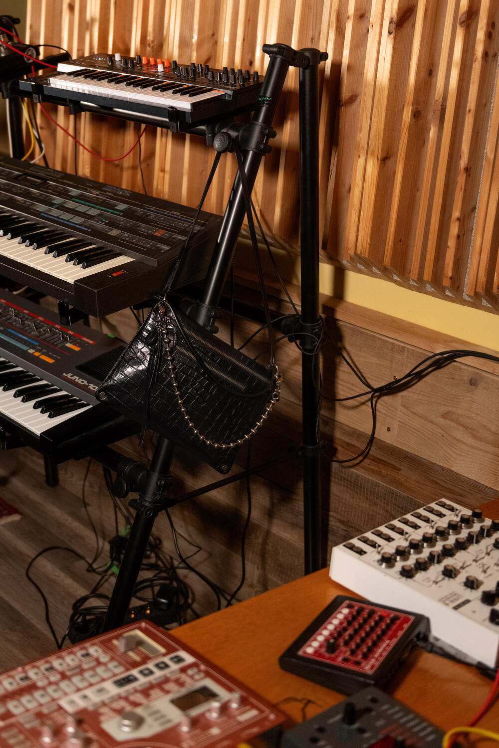 Ein Raum mit Synthesizern auf Ständern, einem Mischpult auf einem Holztisch und einer schwarzen Handtasche von INA KENT mit Kettenriemen, die am Ständer hängt. Der Hintergrund ist mit Holzvertäfelung versehen.