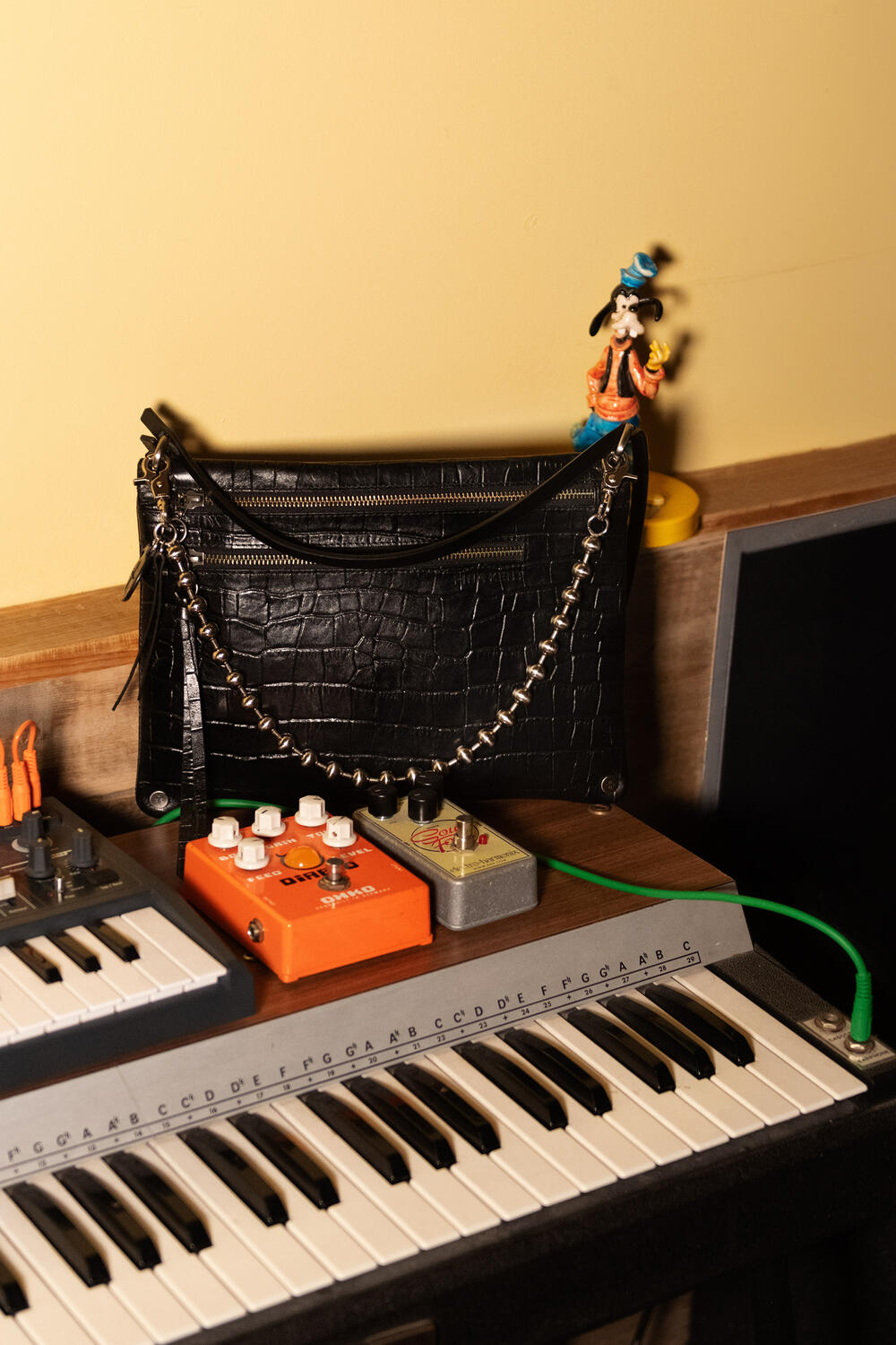 Ein Keyboard und diverse Audiogeräte auf einer Holzoberfläche, dazu eine INA KENT Handtasche und eine Goofy-Figur obendrauf.