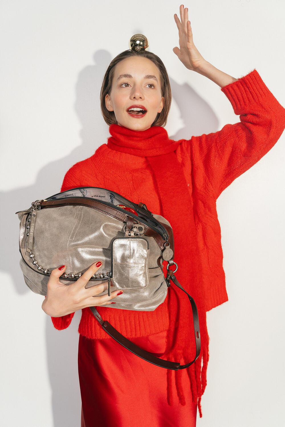 Woman in a red sweater and scarf holds the INA KENT DELA EDEN ed.1 bag in Crackled Anthra leather. She balances a golden Christmas bauble on her head while presenting the bag in front of her upper body.