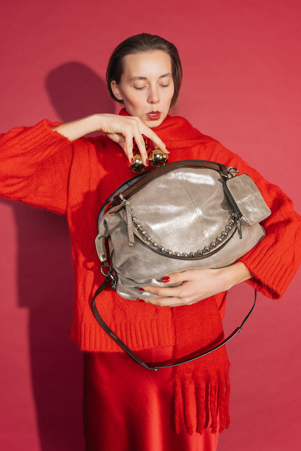 Woman in a red outfit with sweater, scarf and skirt holds the INA KENT bag DELA EDEN ed.1 in Crackled Anthra leather in front of her upper body. She decorates the bag with golden Christmas baubles against a red background.
