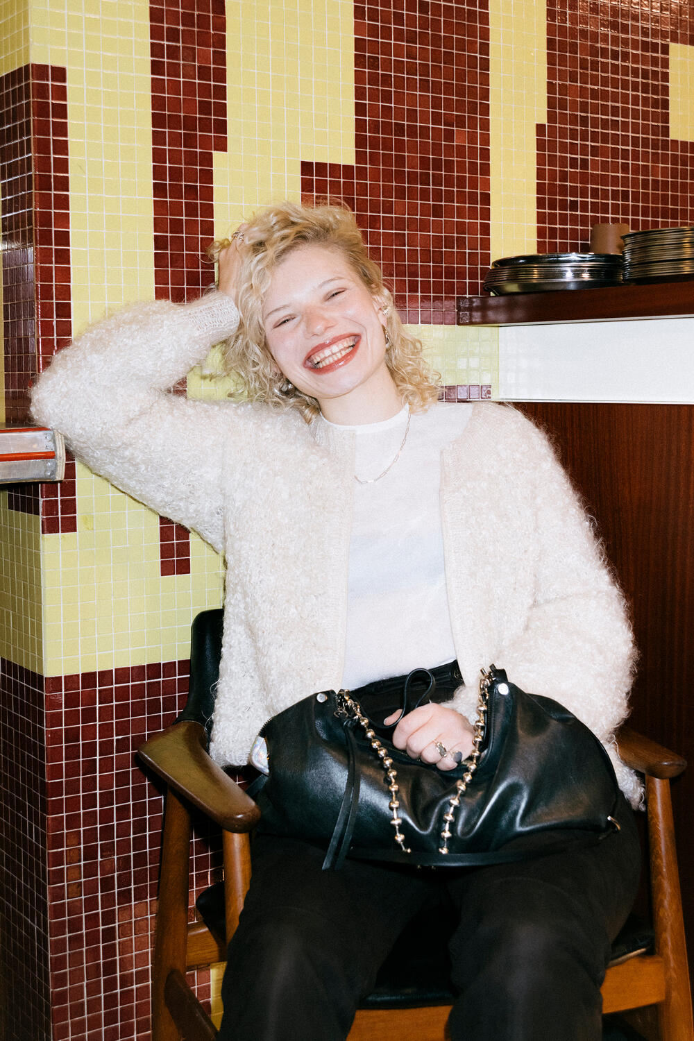  Luisa Gaffron sits in a wooden chair in front of a retro tiled wall, her black INA KENT bag with chain detail casually on her lap as she laughs heartily.