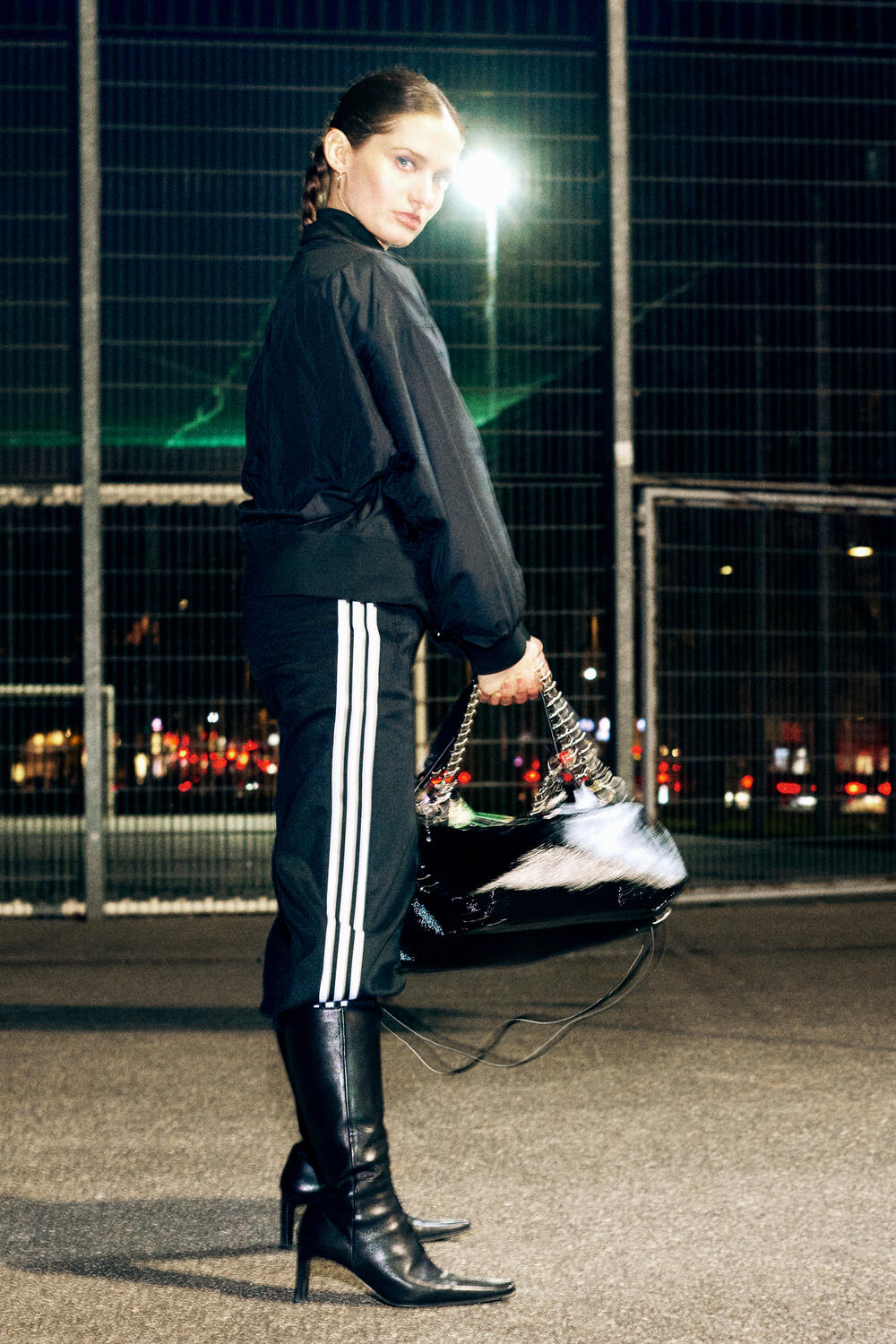 A woman stands outdoors at night in front of a wire fence, wearing a black jacket, black pants with white side stripes, heeled black boots, and holding a large INA KENT black handbag.
