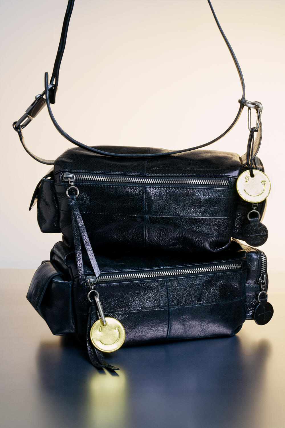 Two black leather INA KENT crossbody bags with front zippers are stacked, each featuring a gold smiley face and round black keychains on a neutral background.