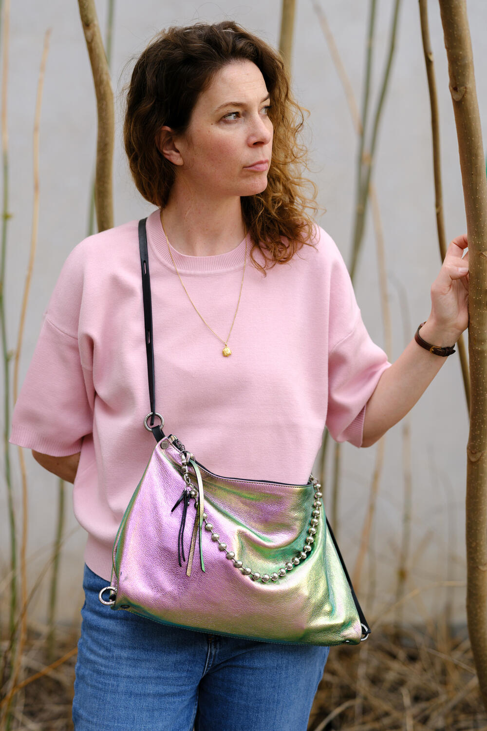 Woman in a pink short-sleeve top and jeans stands outdoors, holding a tree branch. She carries an INA KENT shiny, iridescent handbag with chain and tassel details.