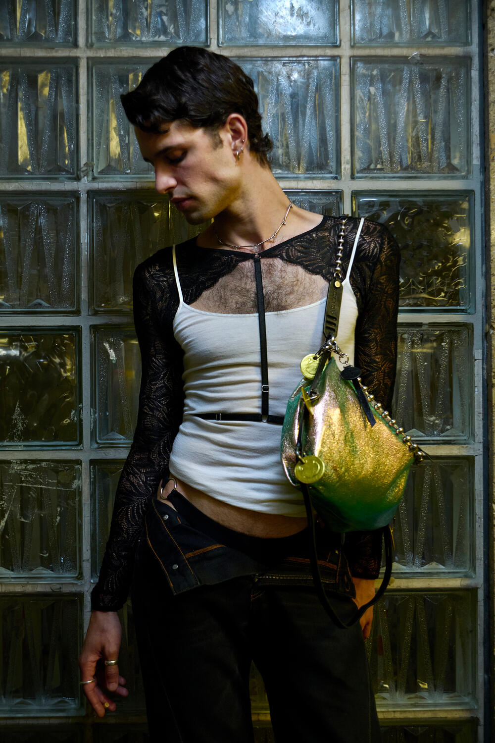 A person wearing a sheer black lace long-sleeve top, white tank, dark pants, and an INA KENT metallic shoulder bag stands in front of glass block windows, looking downward.