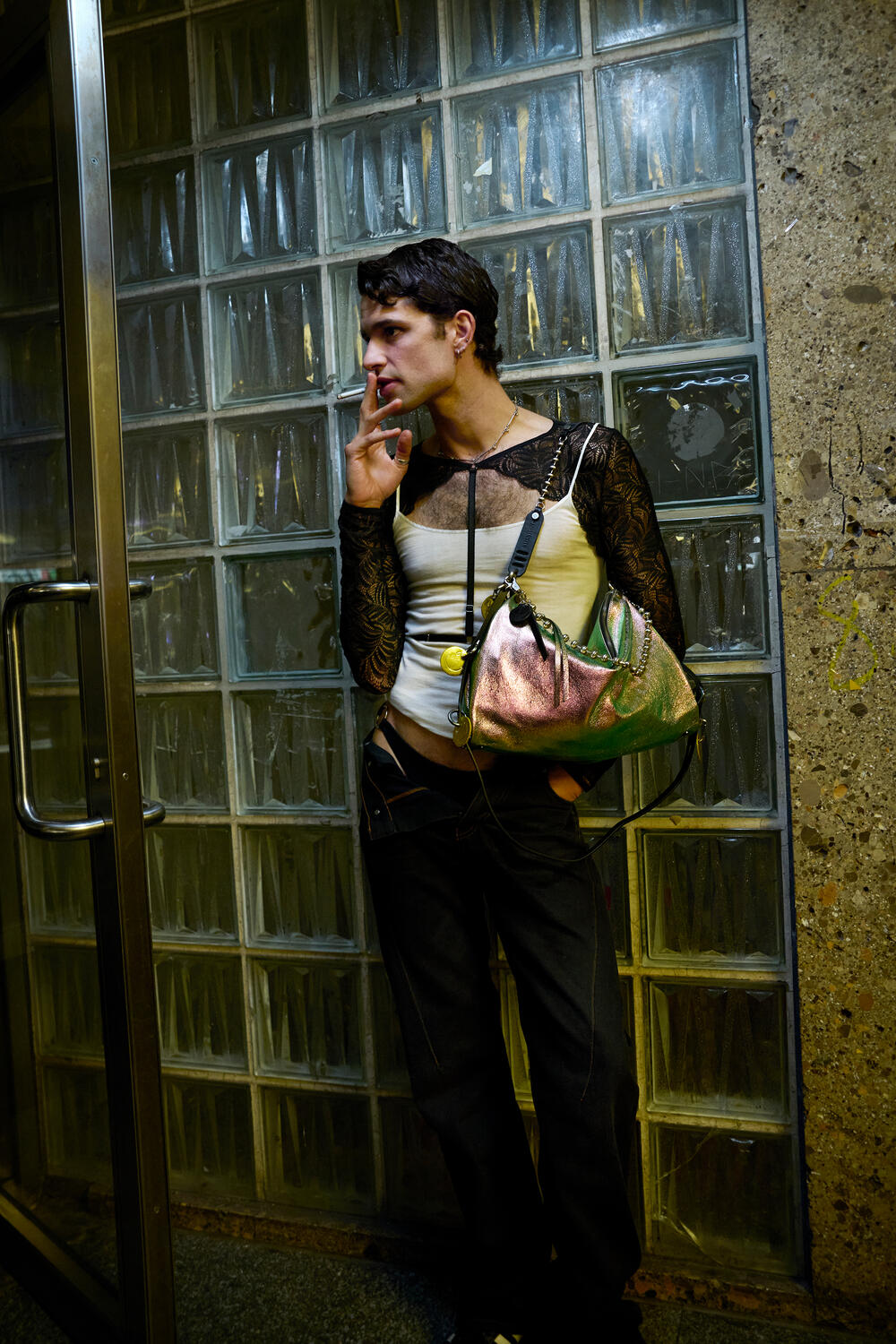 A person stands by a glass block wall and door, holding an INA KENT metallic handbag and smoking a cigarette while gazing into the distance.