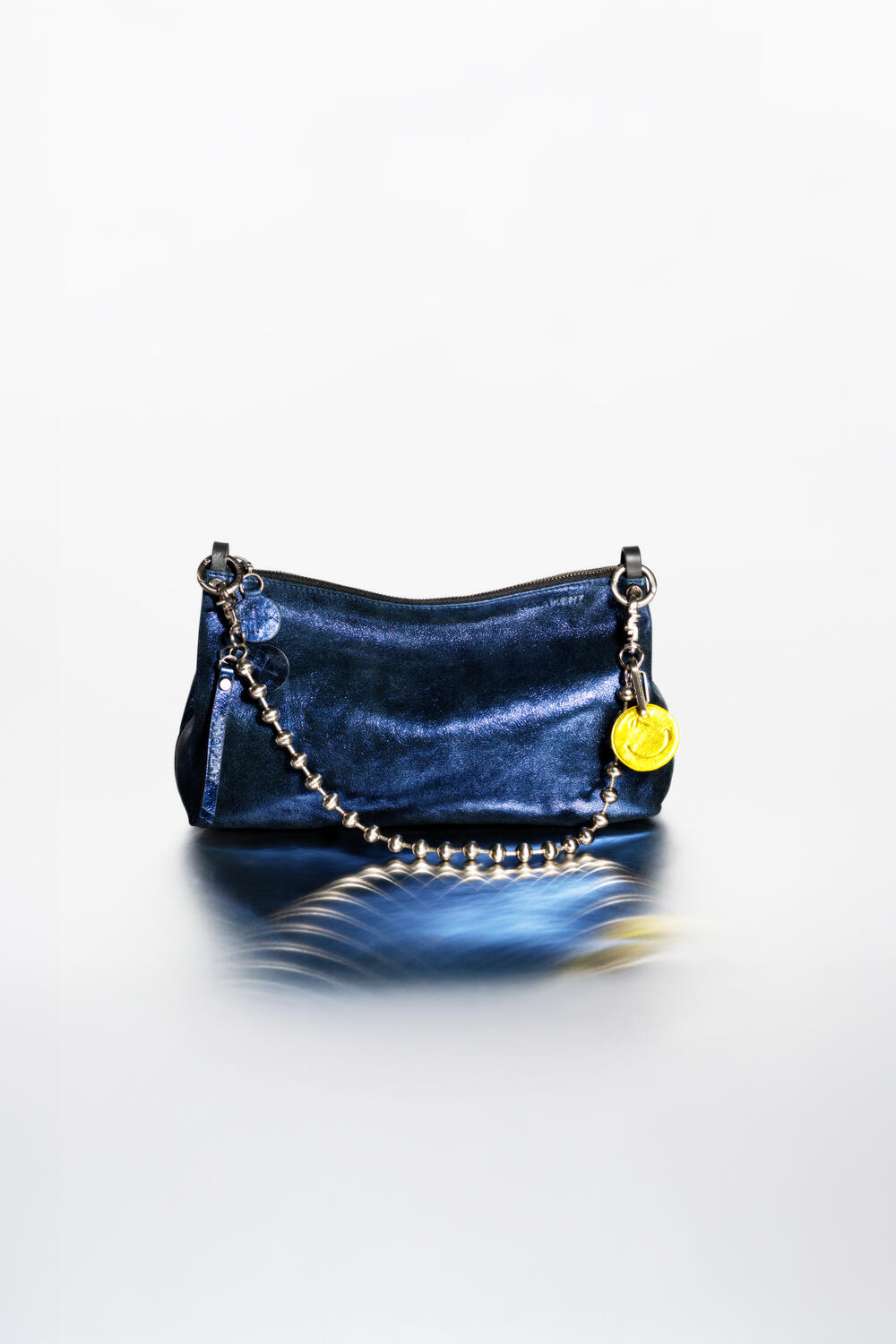 Eine kleine, dunkelblaue INA KENT Handtasche mit einem metallischen Kettenriemen und goldenen und blauen dekorativen Anhängern, dargestellt auf einer reflektierenden Oberfläche mit weißem Hintergrund.
