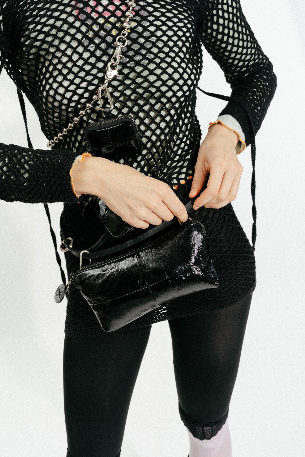 Person, die ein schwarzes Netzoberteil und schwarze Leggings trägt und eine schwarze INA KENT-Ledertasche mit einem Kettenriemen in der Hand hält, vor einem schlichten weißen Hintergrund.