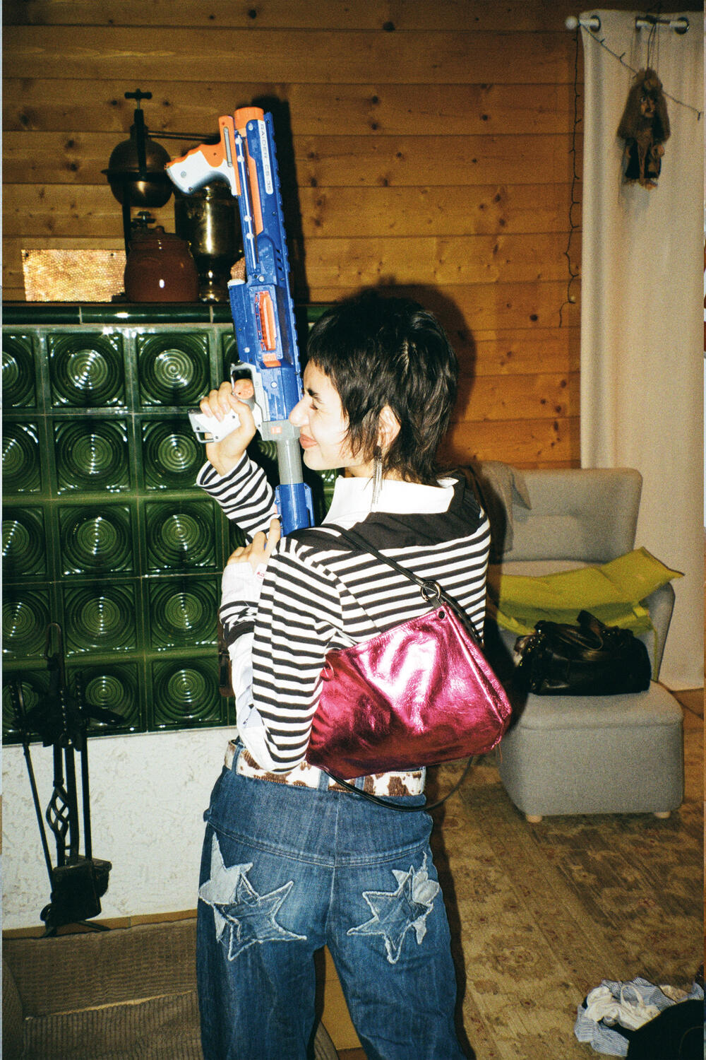 A person in a striped shirt and star-pattern jeans stands indoors, holding a toy gun, with an INA KENT pink bag over their shoulder and wooden-paneled walls in the background.