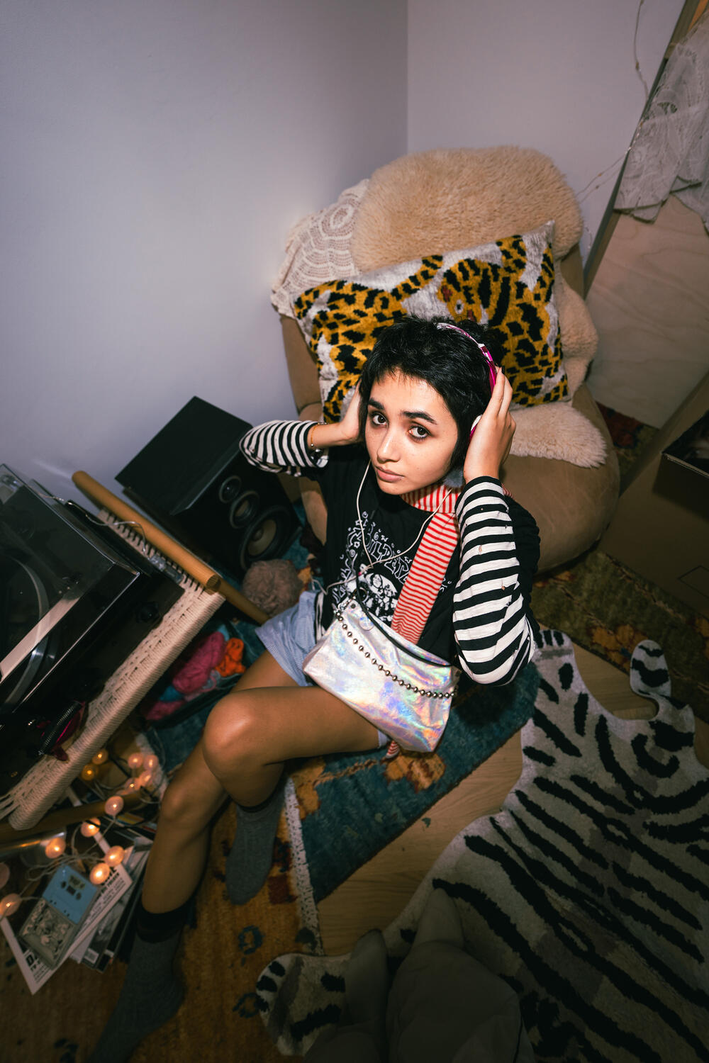A person with short hair and headphones sits in a cluttered room on a cushioned chair, surrounded by candles, a record player, patterned rugs, and an INA KENT bag resting nearby.