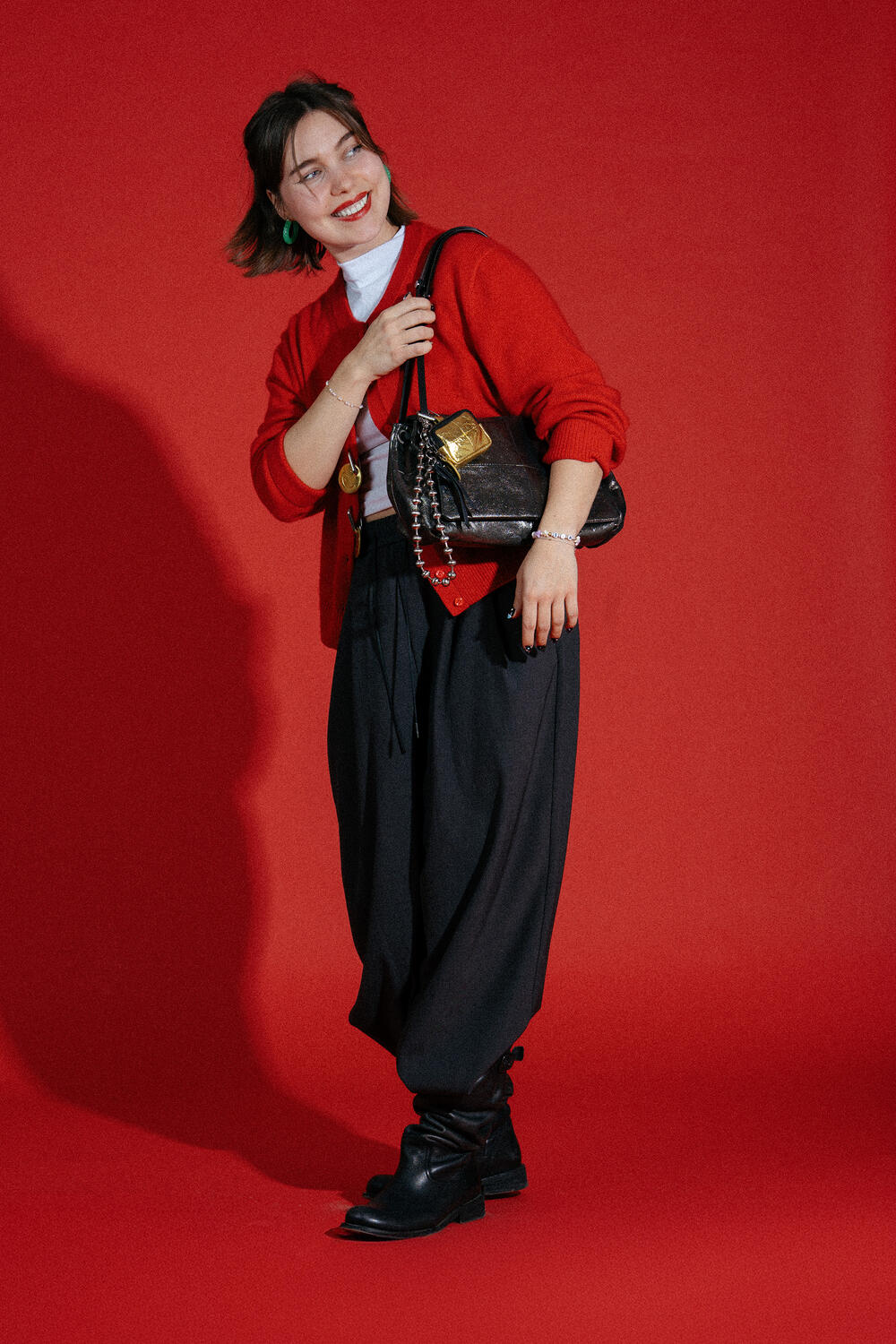 Eine Frau steht vor einem roten Hintergrund, trägt einen roten Pullover, eine schwarze Hose und schwarze Stiefel und hält eine braune Handtasche von INA KENT. Sie lächelt und blickt zur Seite.