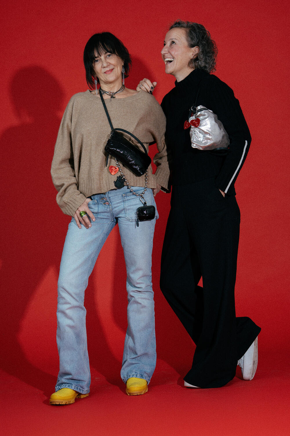 Zwei Frauen stehen vor einem roten Hintergrund und lächeln. Die eine trägt einen beigen Pullover, blaue Jeans und gelbe Schuhe, die andere trägt ein schwarzes Outfit. Beide halten elegante INA KENT-Handtaschen in der Hand.