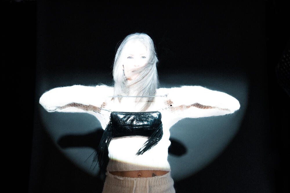 A person with long light hair stands under a bright spotlight, wearing an INA KENT white sweater and a black top, with arms extended horizontally.