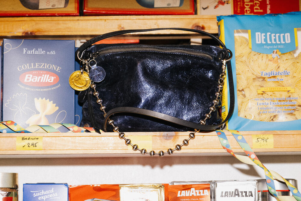 Ein INA KENT Portemonnaie aus schwarzem Leder mit einem Kettenriemen liegt in einem Lebensmittelgeschäft in einem Regal zwischen Barilla Farfalle und De Cecco Tagliatelle.