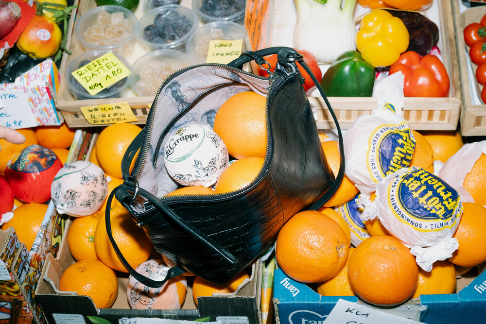 Eine schwarze Handtasche von INA KENT, gefüllt mit Orangen und einem verpackten Käse, liegt auf einem Obst- und Gemüsestand.