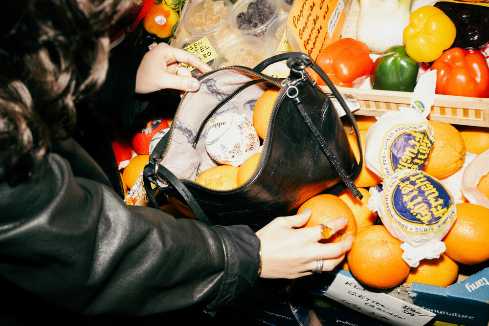 Eine Person legt Orangen aus einer Marktauslage in eine schlichte schwarze INA KENT-Handtasche; anderes Gemüse und verpackte Waren liegen in der Nähe.