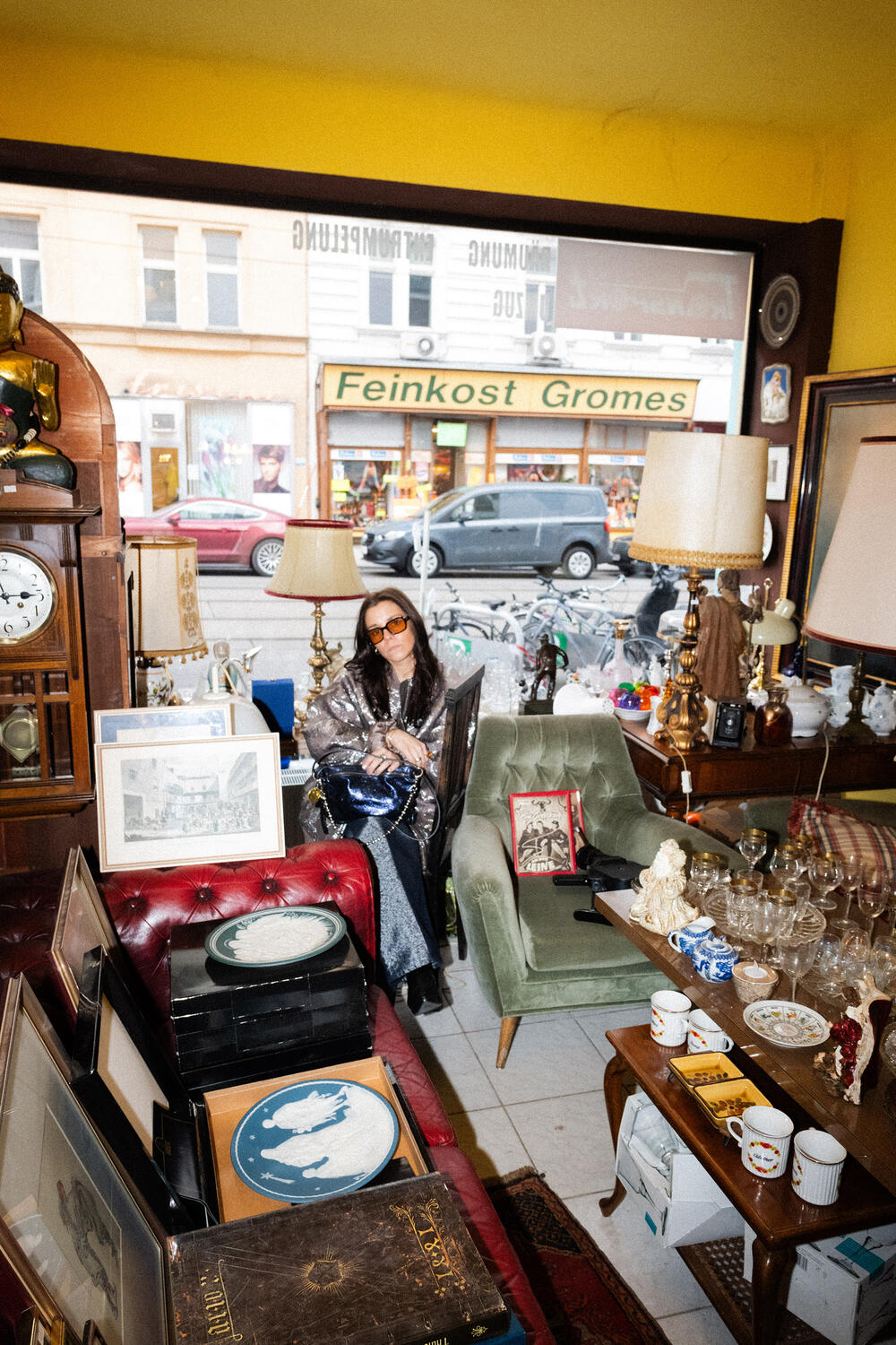 Eine Frau mit Sonnenbrille sitzt in einem Vintage-Laden, der mit Möbeln, Lampen, Glaswaren und gerahmten Bildern gefüllt ist. Ihre INA KENT-Tasche liegt in der Nähe, während das Sonnenlicht durch ein großes Fenster mit Blick auf die Straße fällt.