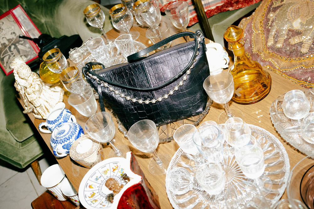 Eine schwarze, strukturierte Handtasche von INA KENT liegt auf einem unordentlichen Holztisch, umgeben von verschiedenen Gläsern, Teetassen, einer goldenen Karaffe und Dekorationsartikeln.