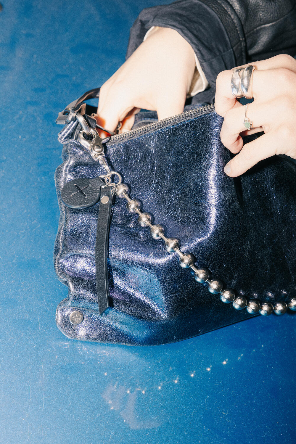 Eine Hand mit silbernen Ringen öffnet eine dunkle, glänzende Lederhandtasche von INA KENT mit einem perlenbesetzten Kettenriemen auf einer blau reflektierenden Oberfläche.