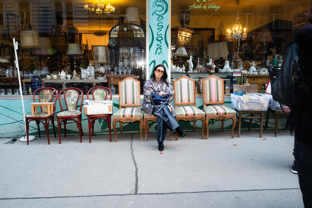 Eine Frau mit Sonnenbrille und einer INA KENT-Tasche sitzt auf einer Reihe von Vintage-Stühlen vor einem Antiquitätengeschäft, in dessen Schaufenster und auf dem Gehweg verschiedene Gegenstände ausgestellt sind.