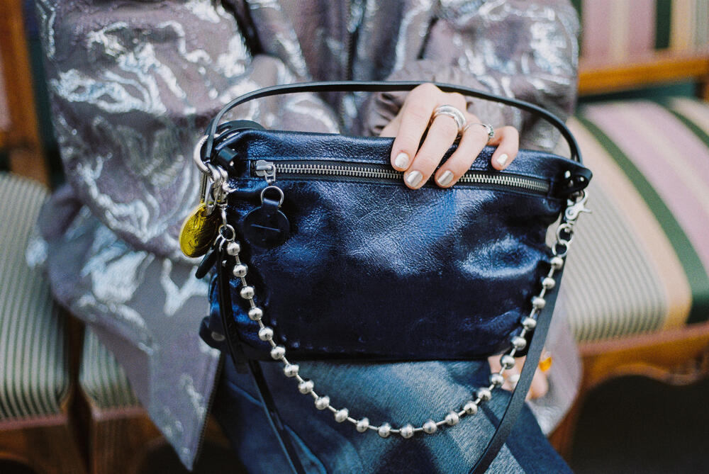 Eine Person, die eine gemusterte silberne Jacke trägt, hält eine glänzende schwarze Handtasche von INA KENT mit einem perlenbesetzten Riemen und einem Metallreißverschluss, während sie auf einem gestreiften Stuhl sitzt.