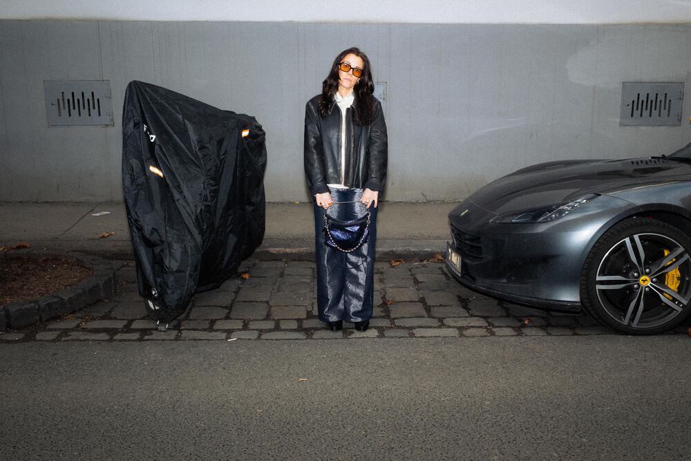 Eine Frau mit Sonnenbrille, schwarzer Lederjacke und weiten Hosenbeinen steht auf einem Bürgersteig zwischen einem überdachten Fahrzeug und einem grauen Sportwagen und trägt eine INA KENT-Tasche.