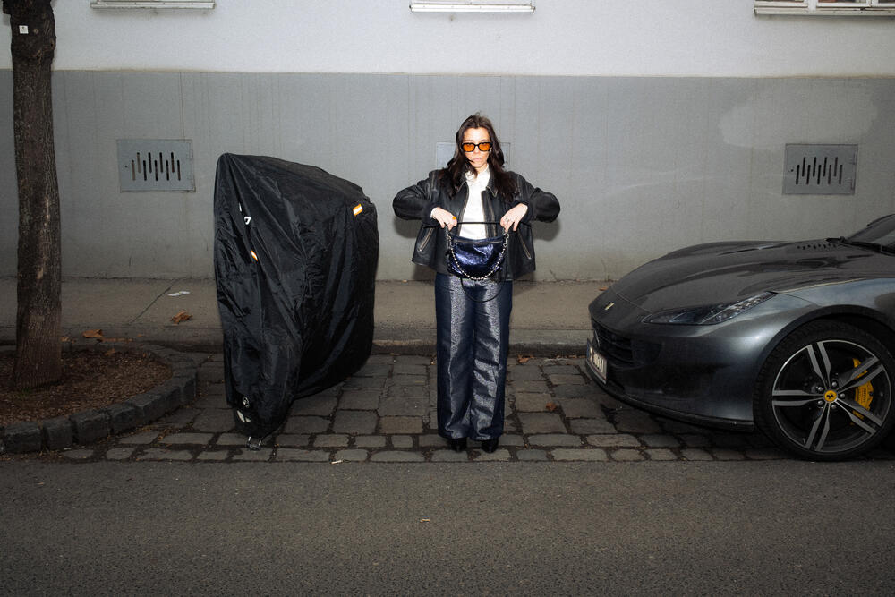 Eine Person mit Sonnenbrille, Lederjacke und weiter Hose steht auf einem kopfsteingepflasterten Gehweg zwischen einem überdachten Objekt und einem geparkten Sportwagen und hält stilvoll eine offene INA KENT-Handtasche in der Hand.