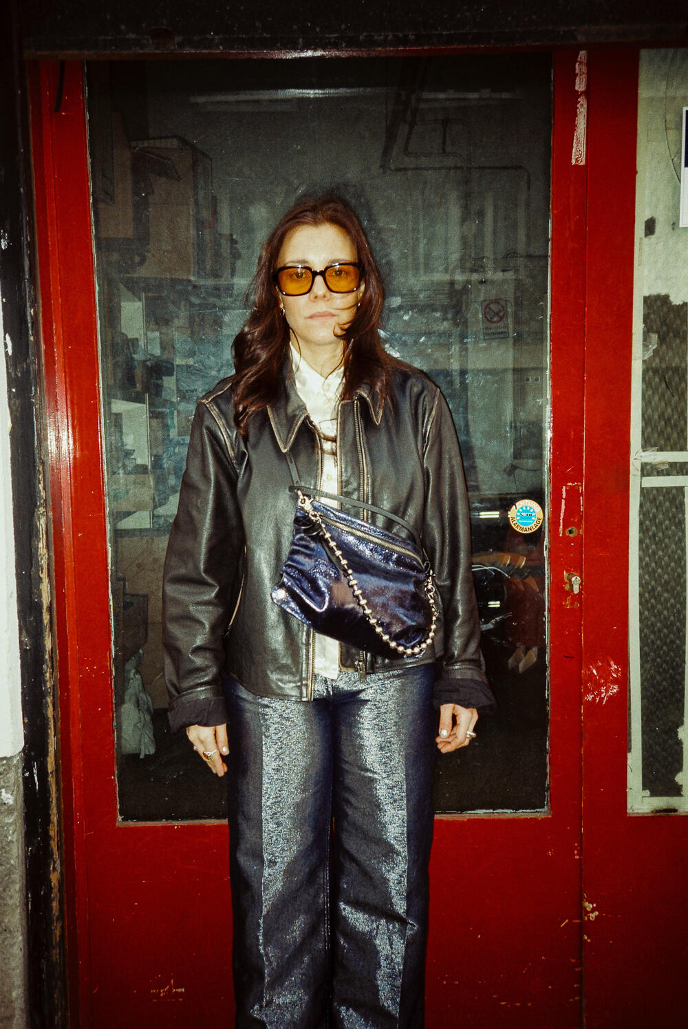 Eine Frau mit Sonnenbrille, dunkler Lederjacke und glänzender Hose steht vor einer rot gerahmten Glastür und hält eine dunkle INA KENT Crossbody Bag in der Hand.