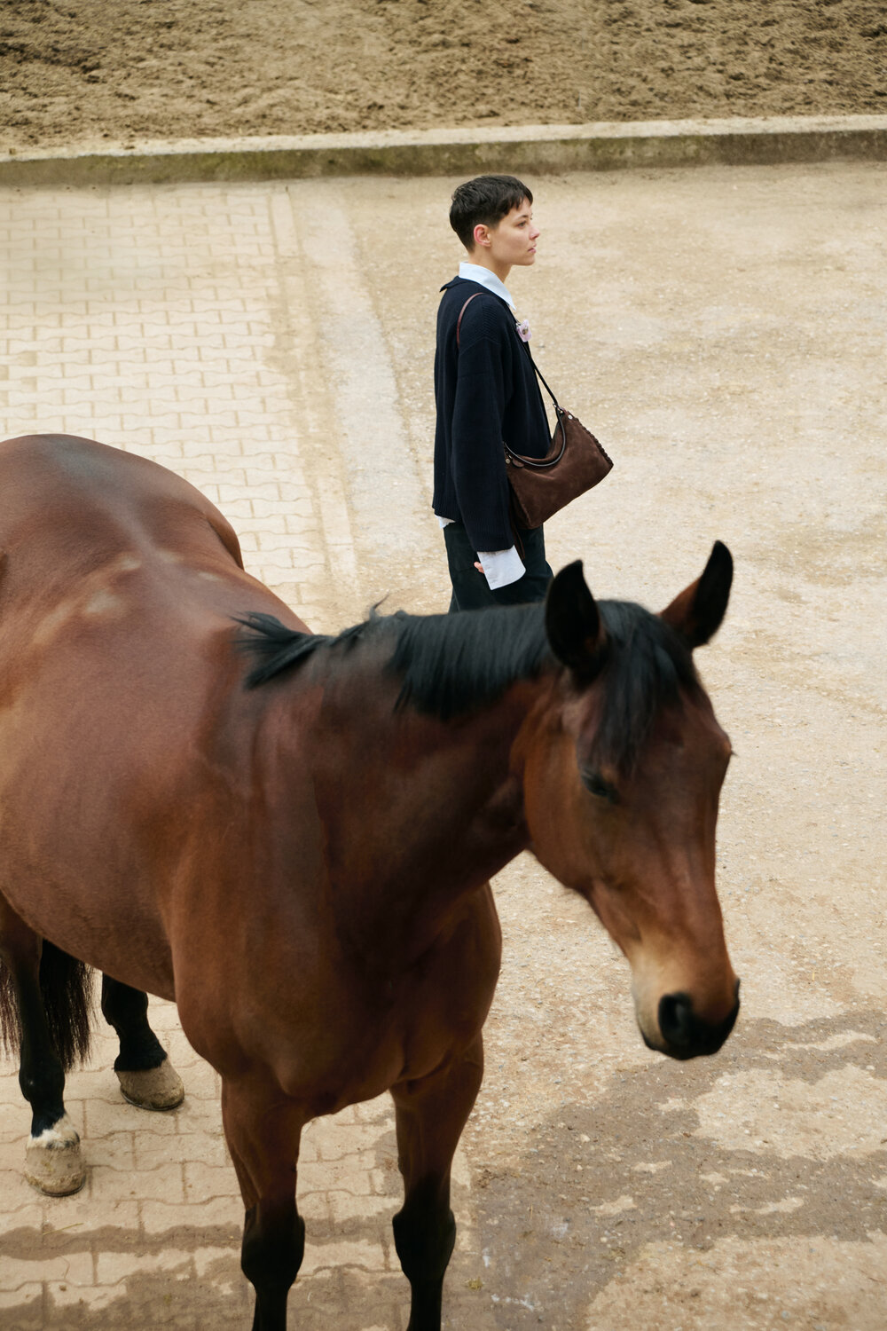 A person standing on a paved area next to a brown horse in an outdoor setting, carrying an INA KENT bag.