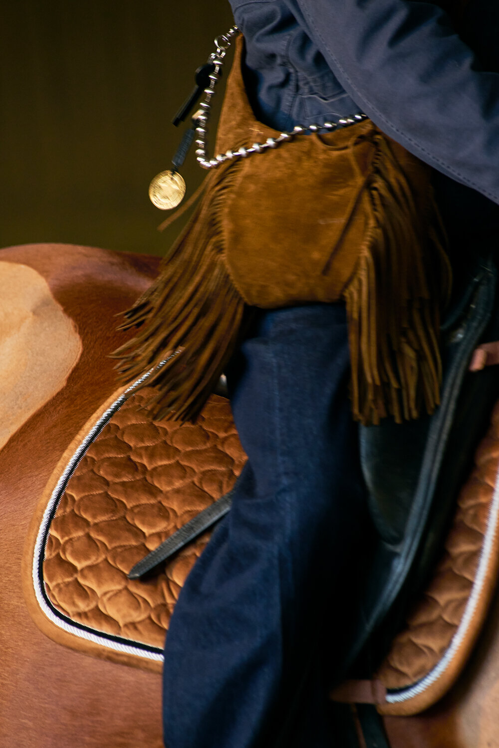 A person wearing blue jeans and an INA KENT brown fringed bag is sitting on a brown quilted saddle on a horse.