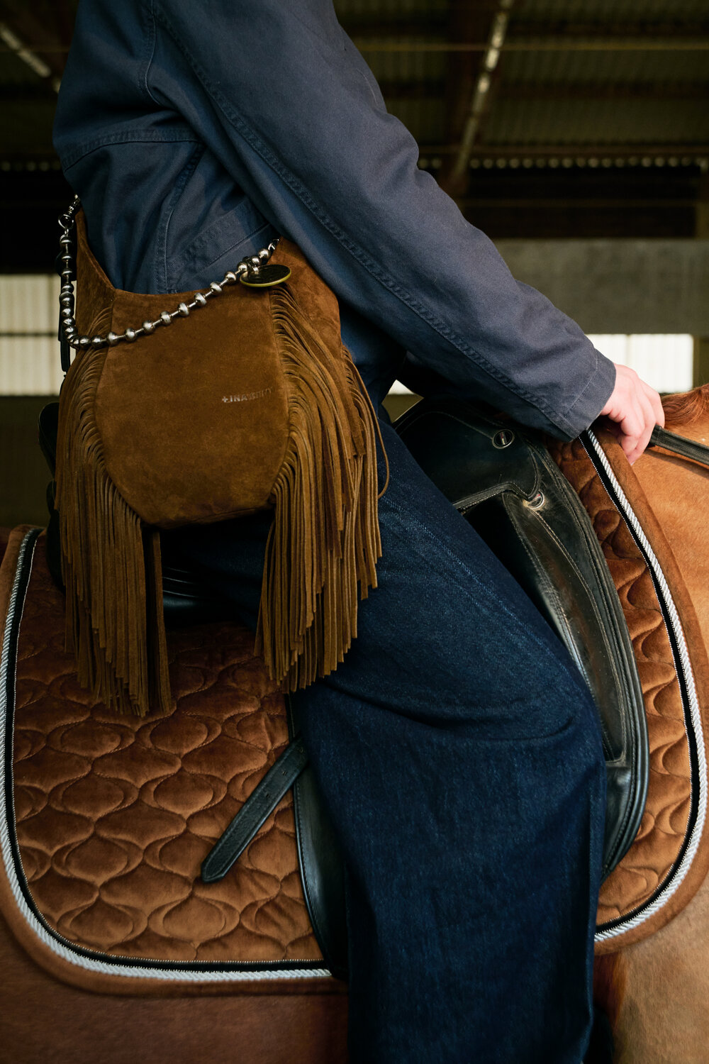 A person in a dark jacket and jeans sits on a saddled brown horse, carrying an INA KENT brown suede fringe bag with a chain strap.