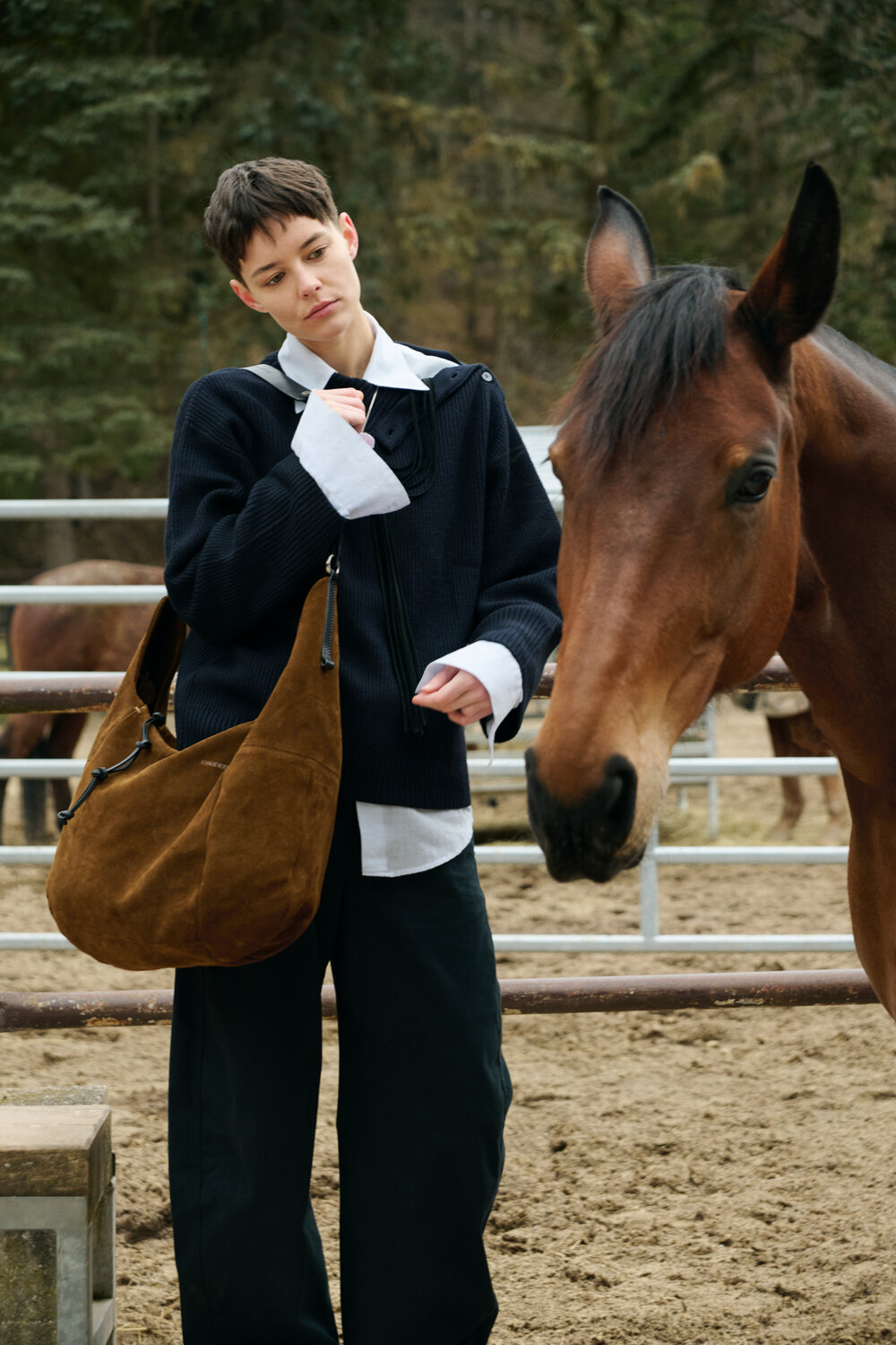 A person with short hair and an INA KENT brown shoulder bag stands next to a brown horse in an outdoor fenced area.