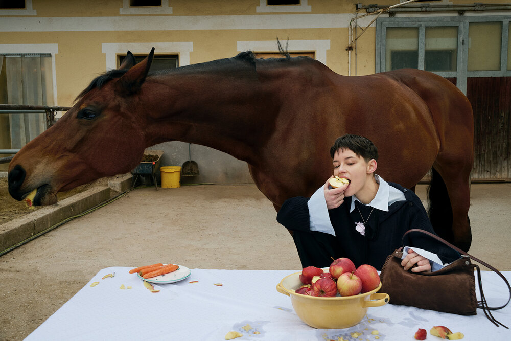 A person sits at a table eating an apple from an INA KENT bag, while a horse beside them also eats an apple. A bowl of apples and some carrots are on the table.