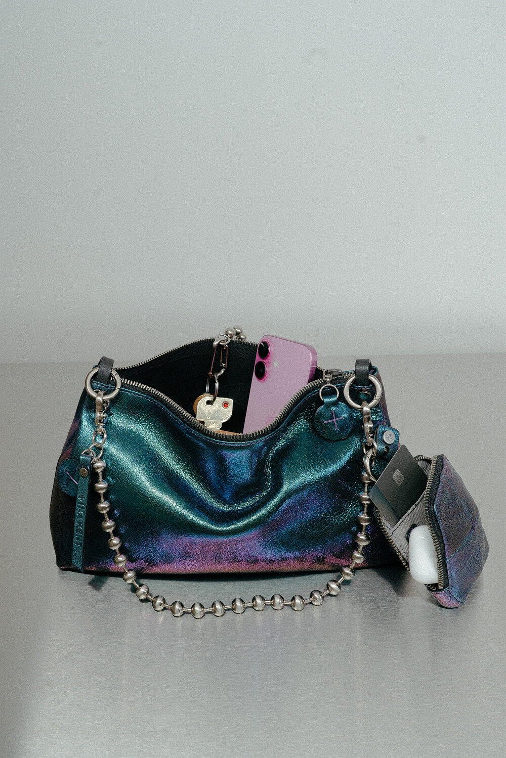 Eine metallische blau-grüne INA KENT Handtasche mit einem perlenbesetzten Kettenriemen, die ein pinkfarbenes Telefon, Schlüssel und eine kleine Tasche mit kabellosen Ohrhörern enthält, steht auf einer grauen Fläche.