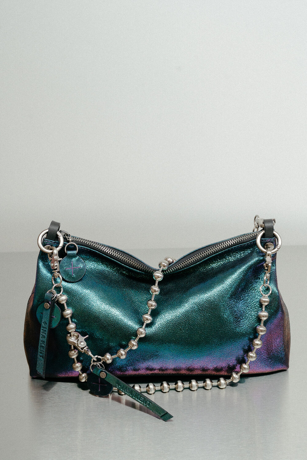 Schillernde Metallic-Handtasche von INA KENT mit Reißverschluss, silbernem Kugelkettenriemen und passenden Reißverschlussanhängern, dargestellt auf einer glatten Oberfläche vor einem neutralen Hintergrund.