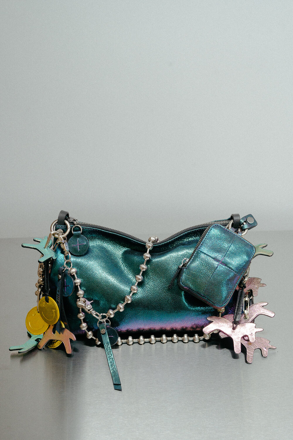 Eine metallisch tealfarbene INA KENT Handtasche mit einem kleinen passenden Beutel, verziert mit einer silbernen Kugelkette und verschiedenen bunten abstrakten Anhängern.