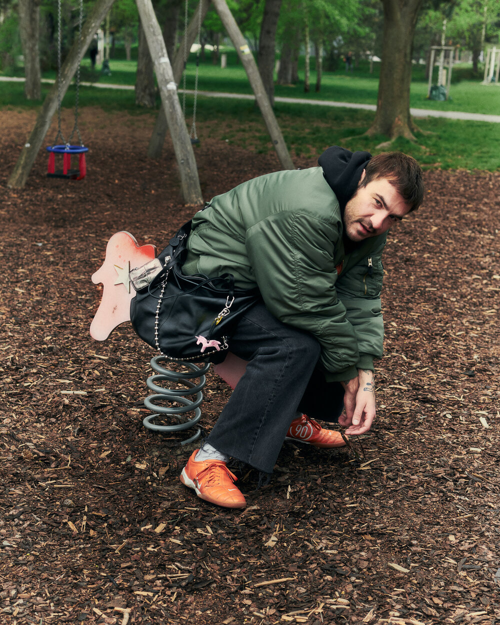 Ein Mann in grüner Jacke und orangefarbenen Schuhen sitzt auf einem rosa Frühlingsreiter auf einem Spielplatz, umgeben von Holzspänen und Bäumen, seine INA KENT-Tasche liegt neben ihm.