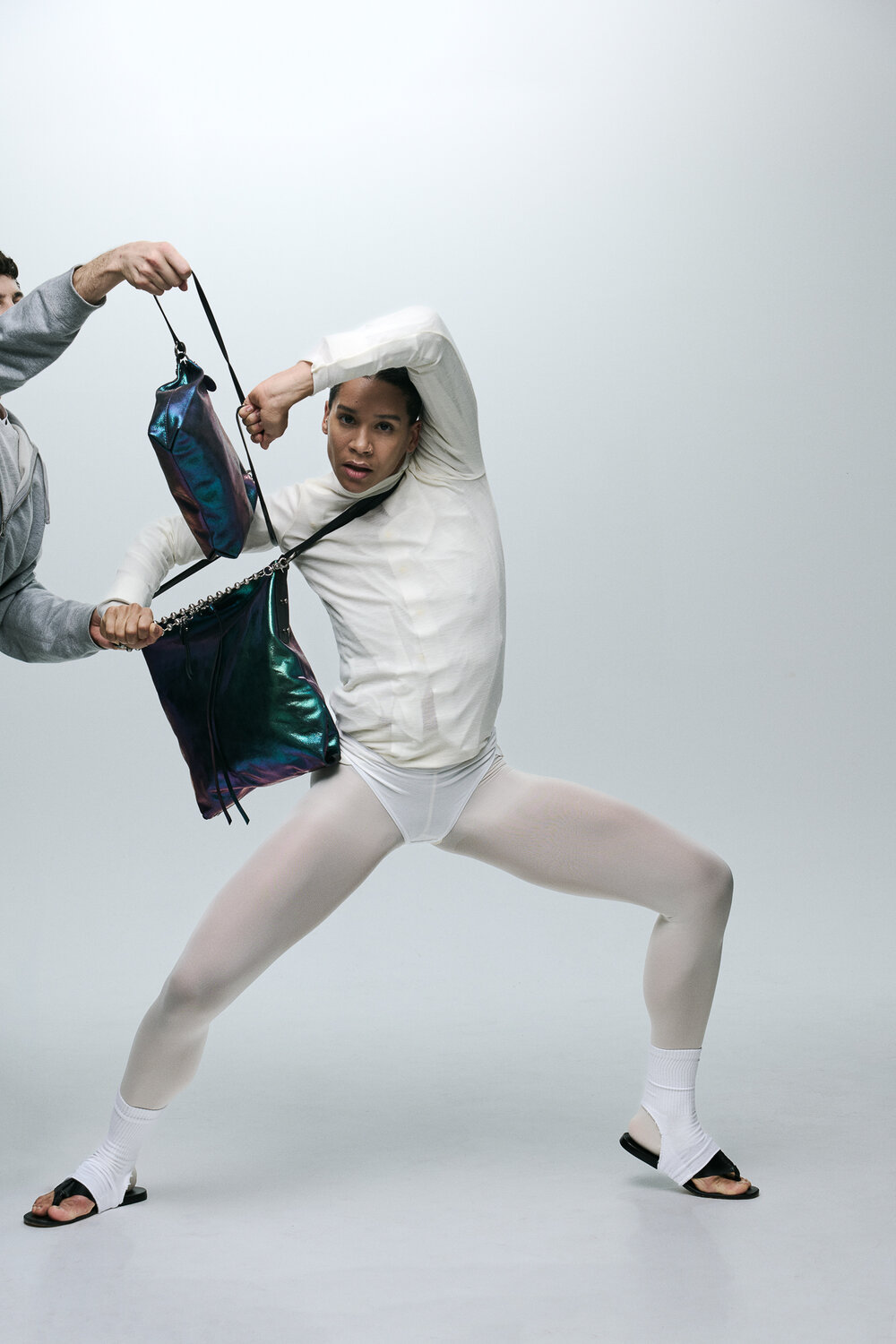 Eine Person in weißer Tanzkleidung hält eine glänzende INA KENT-Tasche, während eine andere Hand versucht, die Tasche wegzuziehen, vor einem schlichten Hintergrund.