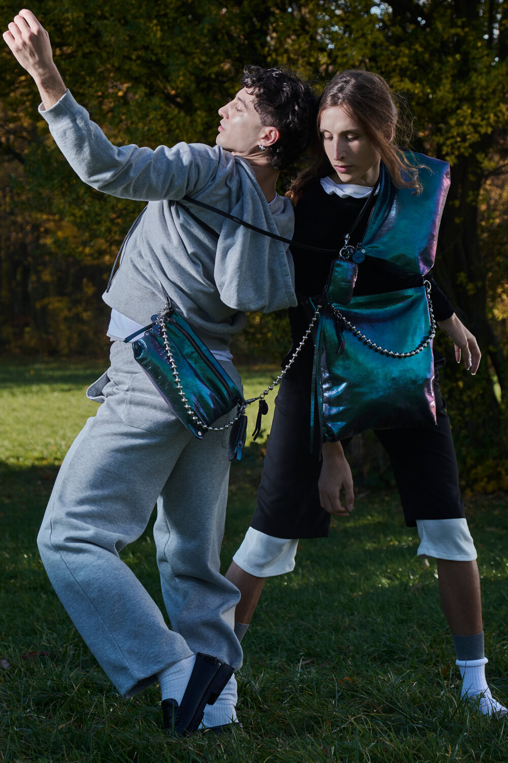 Zwei Personen posieren im Freien in futuristischen Outfits mit INA KENT-Crossbody-Taschen aus Metall und mehrlagiger Kleidung, die auf einer Wiese mit Bäumen im Hintergrund stehen.