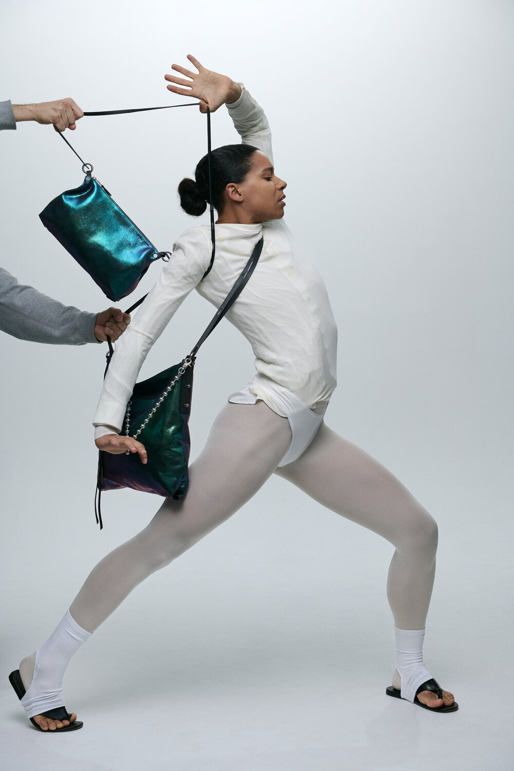 Eine Person in weißer Kleidung und Leggings posiert dynamisch, während jemand aus dem Off zwei blaugrüne INA KENT-Taschen über Schulter und Arm vor einem schlichten Hintergrund präsentiert.