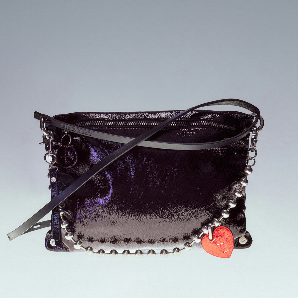 A black patent leather INA KENT handbag with a silver chain detail and a red heart-shaped charm, displayed against a light gradient background.