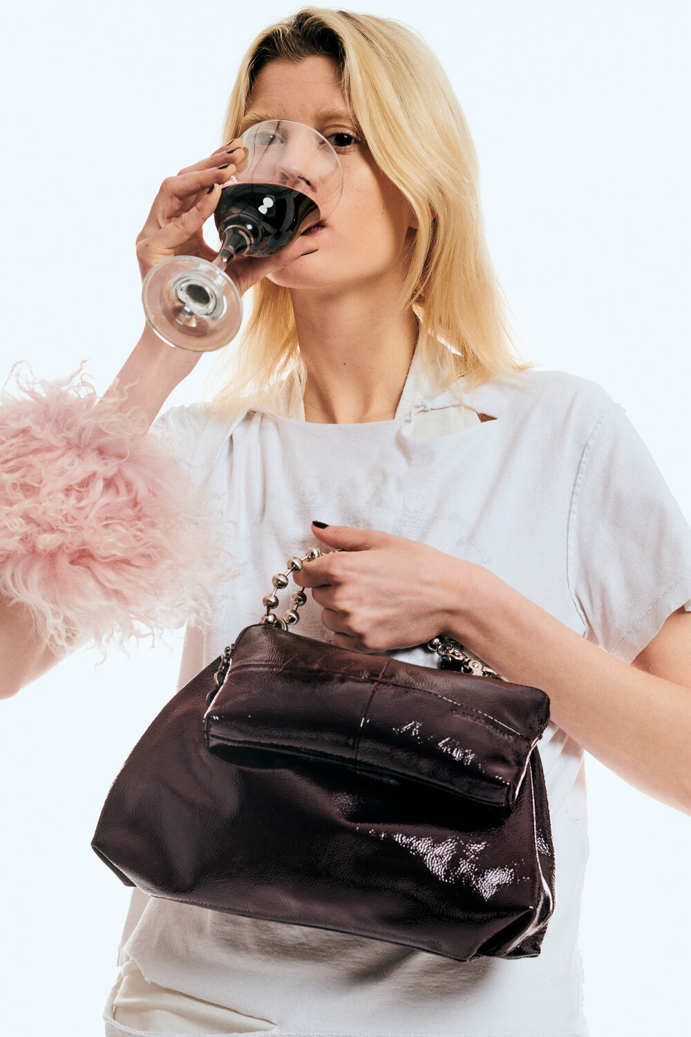A woman in a white shirt holds a large, shiny dark INA KENT handbag and drinks from a wine glass. She wears a pink feathery accessory on her sleeve.