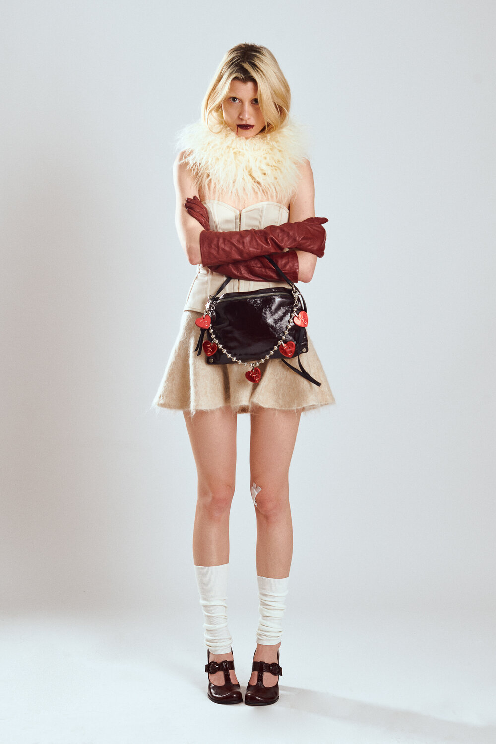 A woman stands against a plain background wearing a fluffy white collar, cream mini skirt, long brown gloves, white socks, brown heels, and an INA KENT black handbag adorned with red charms.
