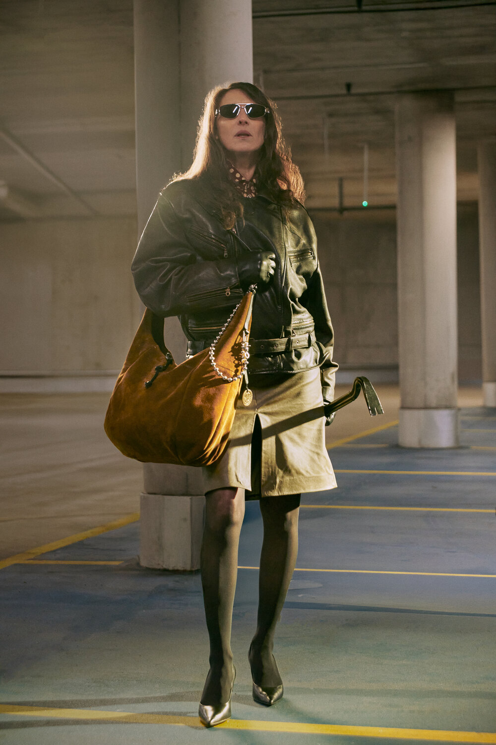 Eine Person mit Sonnenbrille, schwarzer Lederjacke, Handschuhen, Rock und hochhackigen Schuhen steht in einem größtenteils leeren Parkhaus, in der Hand eine große braune INA KENT-Tasche und einen schwarzen Stock.