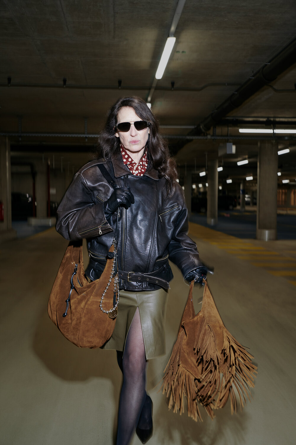 Eine Frau mit Sonnenbrille, schwarzer Lederjacke und grünem Rock geht mit zwei braunen INA KENT-Fransentaschen durch eine Tiefgarage.