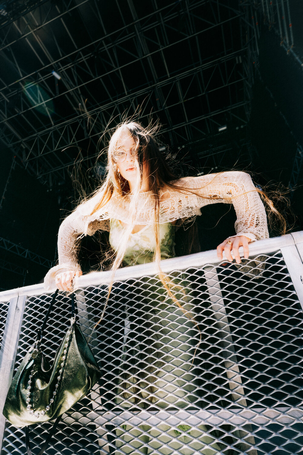 A person with long hair and glasses leans over a metal fence, with an INA KENT green handbag hanging on the fence and stage equipment visible in the background.