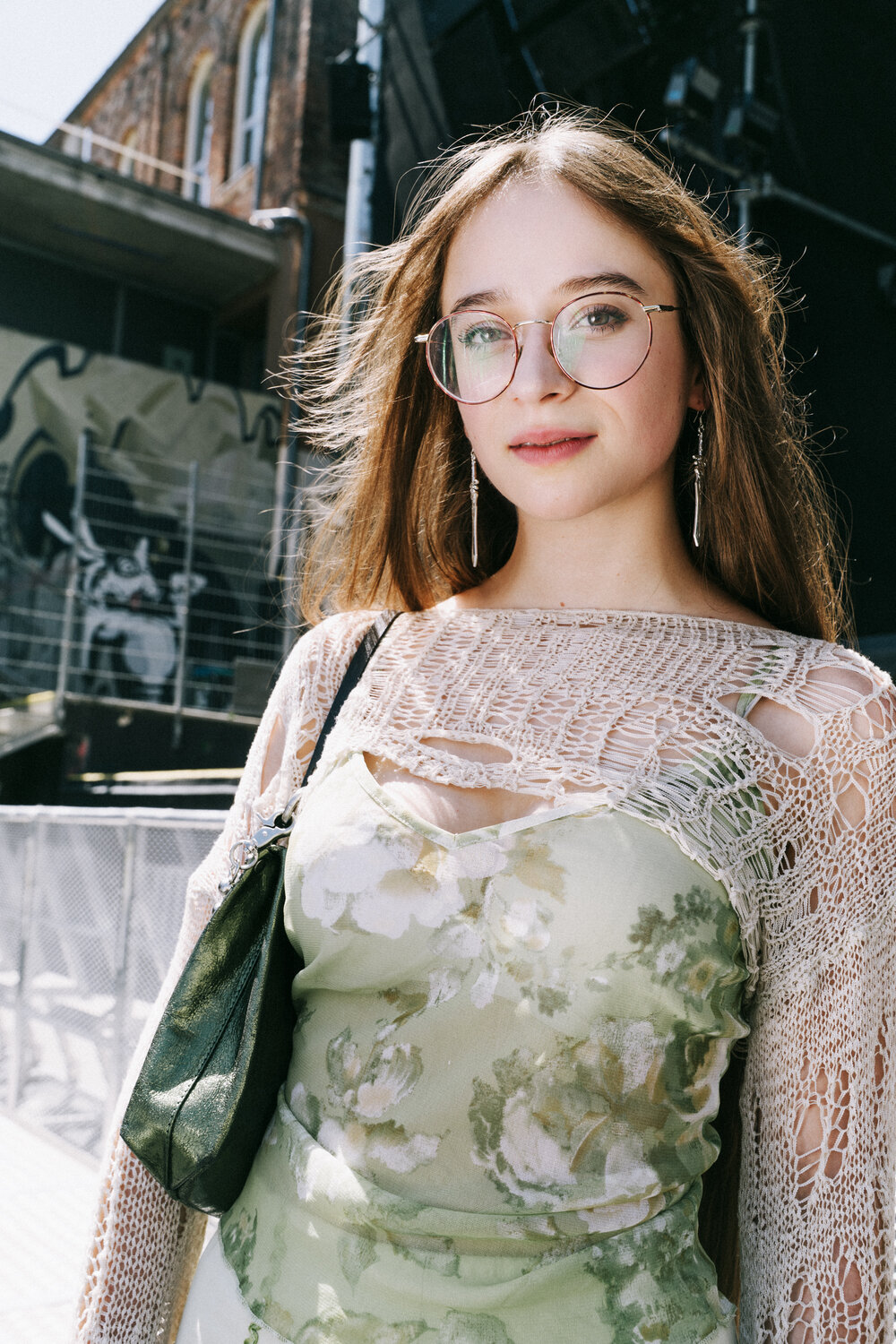 A woman wearing glasses, a green floral dress, an INA KENT bag, and a crocheted shawl stands outdoors in sunlight, with a building and mural in the background.
