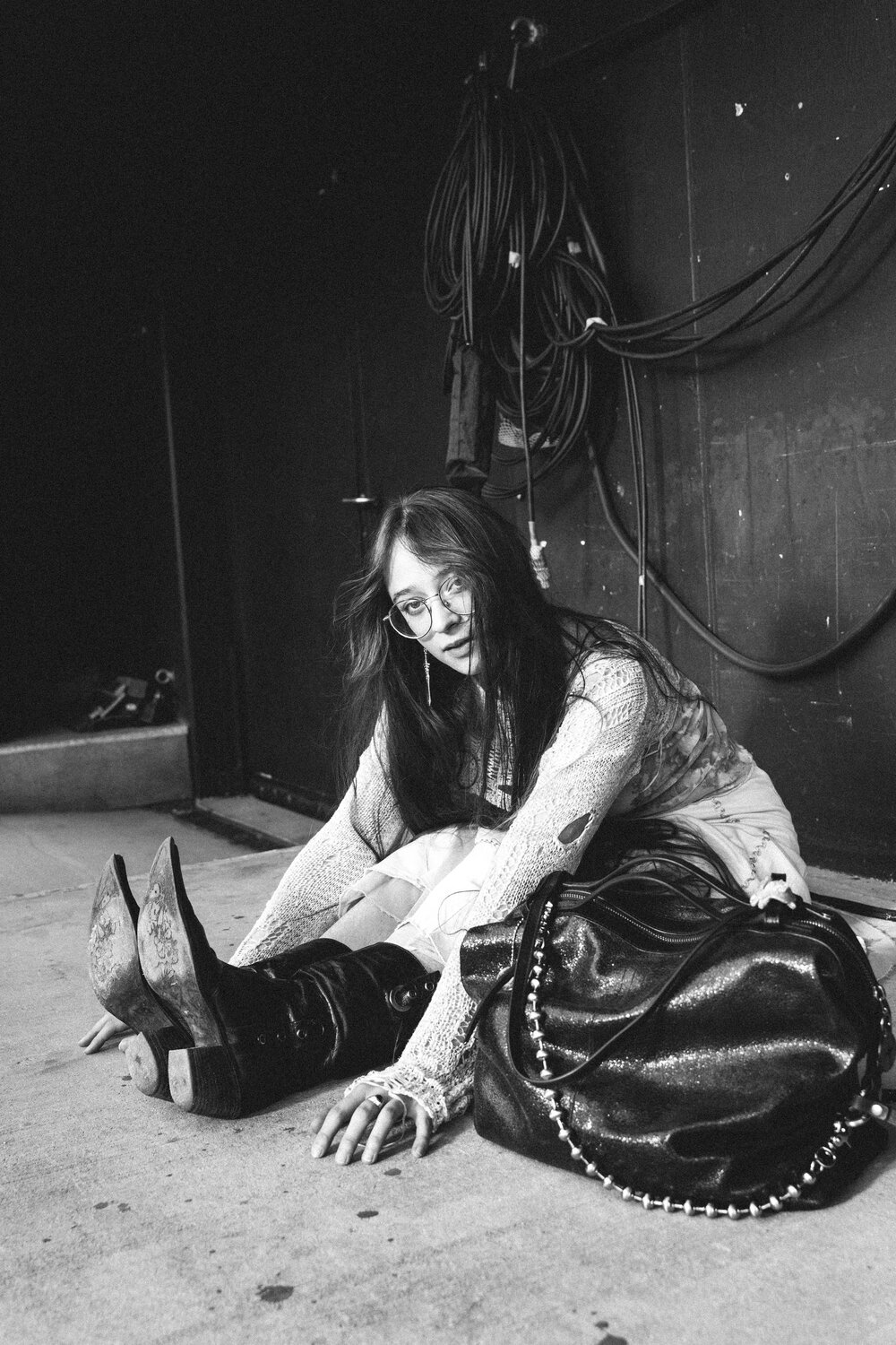 A woman wearing glasses and boots sits on the floor, stretching her legs forward, with an INA KENT bag beside her and cables hanging on the wall in the background.