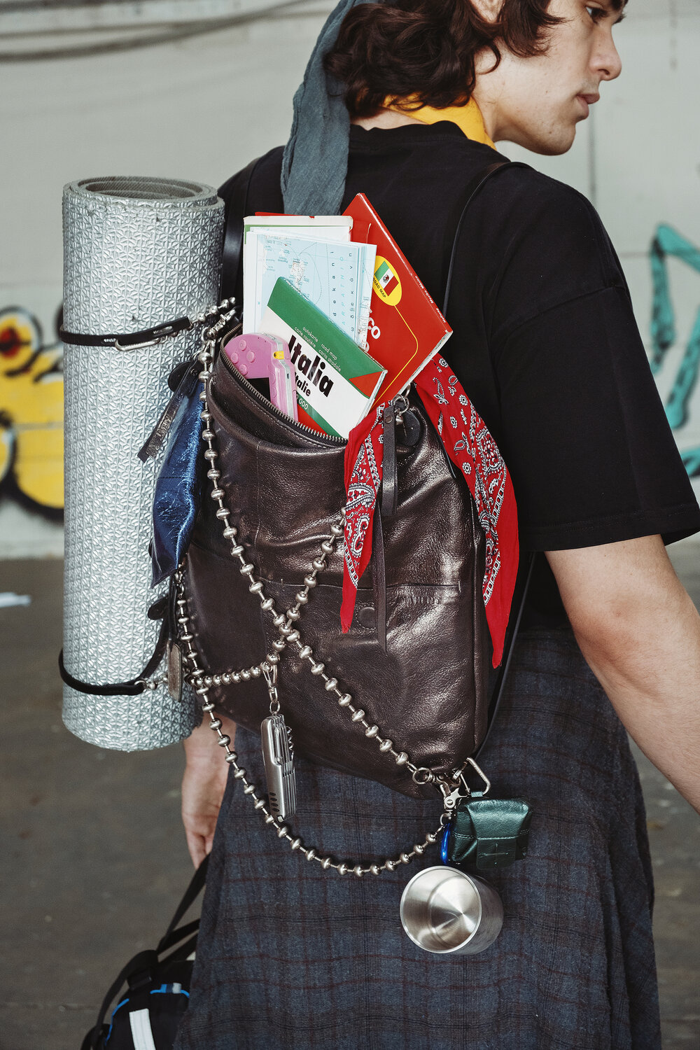 Person wearing a black t-shirt carries an INA KENT backpack with a rolled mat, travel guides, a bandana, and various items attached, including a cup, a chain, and dog tags.